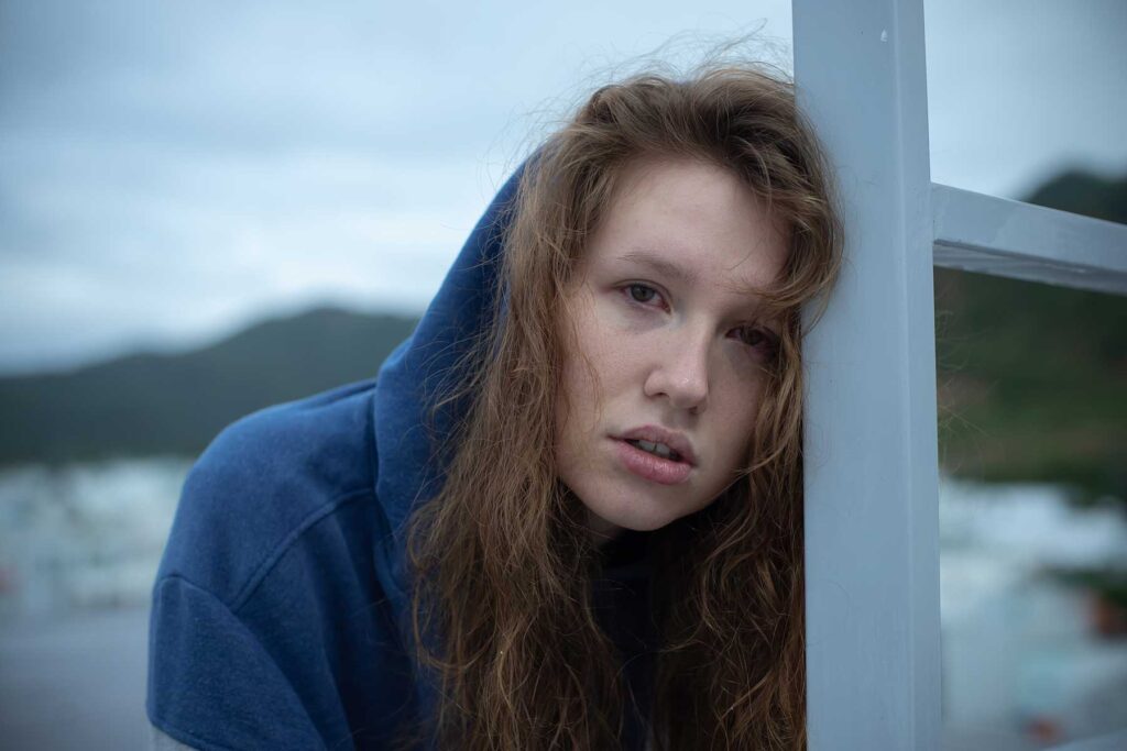 a person with unruly hair leans against a pole outside and looks exhausted while showing signs of cocaine use