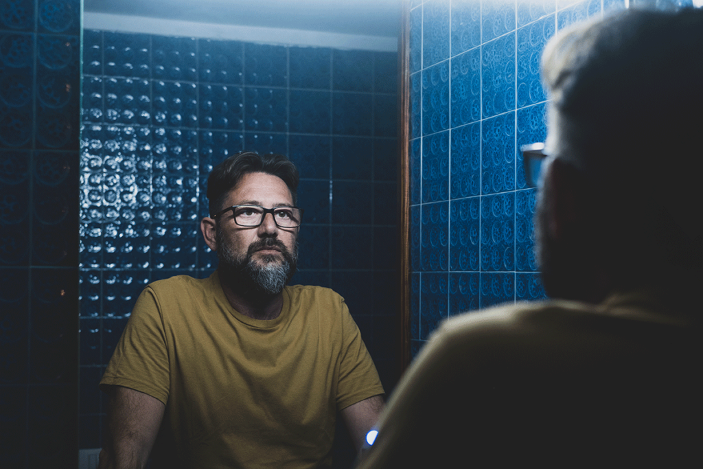 a man with glasses looks at his reflection in a mirror and thinks about the signs of painkiller abuse