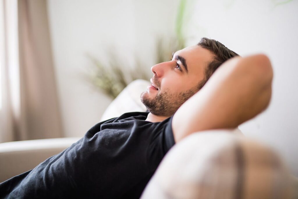 man leans back while sitting on his couch and arms behind his head while thinking about what is cbd