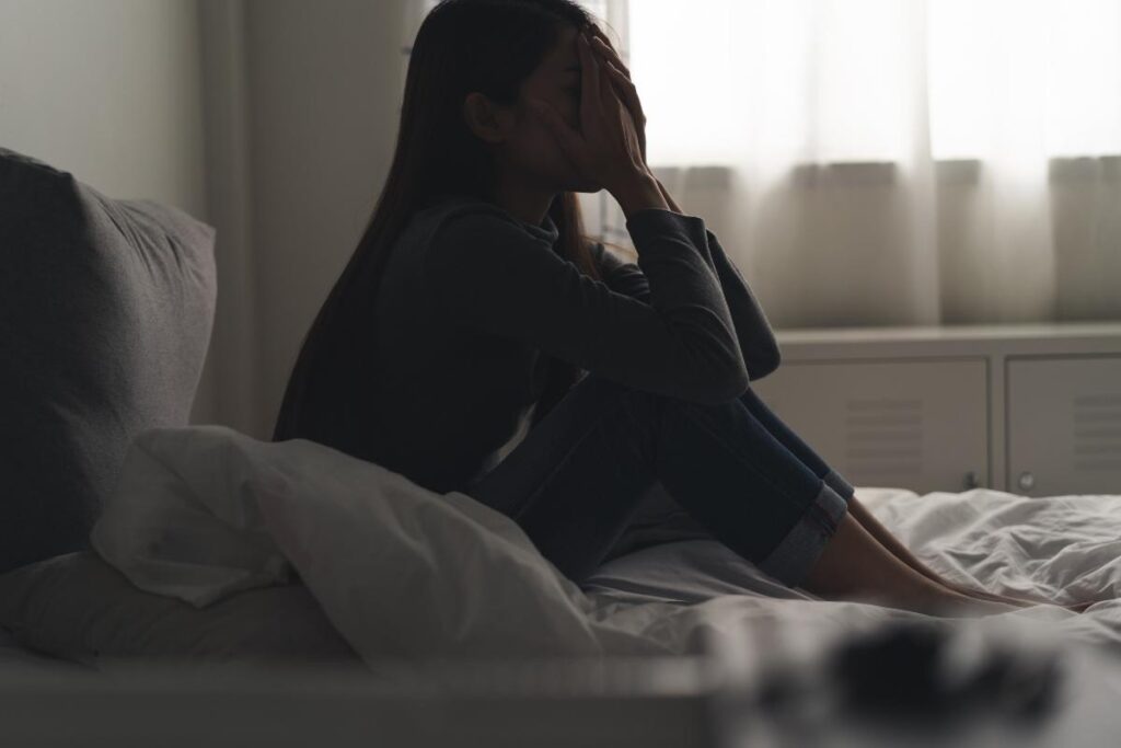 A woman sits on her bed with her knees pulled to her chest. She is experiencing the effects of marijuana and depression.